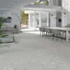 Bright open-plan living and dining area with terrazzo floor and white-gray furniture, opening to a patios through large glass doors far wall light-filled exterior.